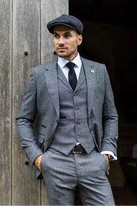 Hombre con traje y gorra grises de pie contra una pared de madera.