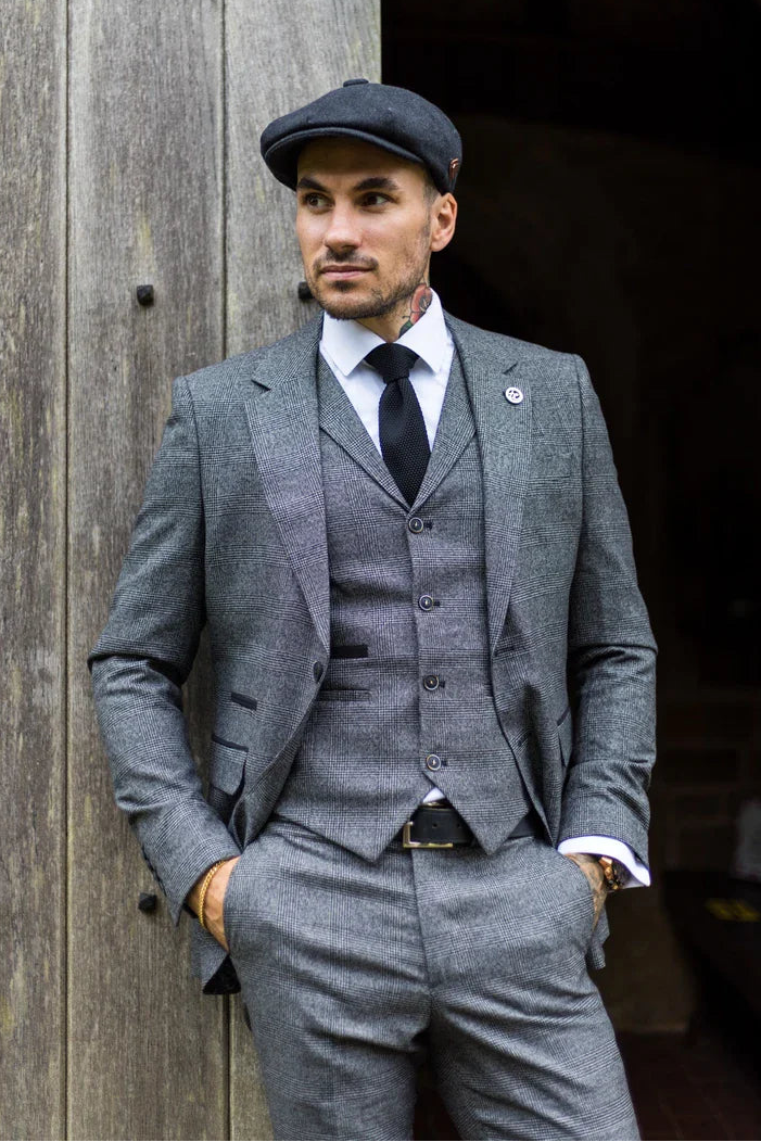 Hombre con traje y gorra grises de pie contra una pared de madera.