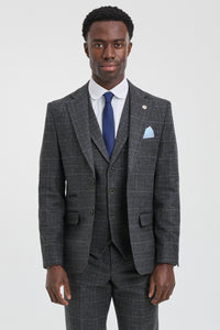 Hombre con traje gris a cuadros, corbata azul y pañuelo de bolsillo sobre fondo blanco.