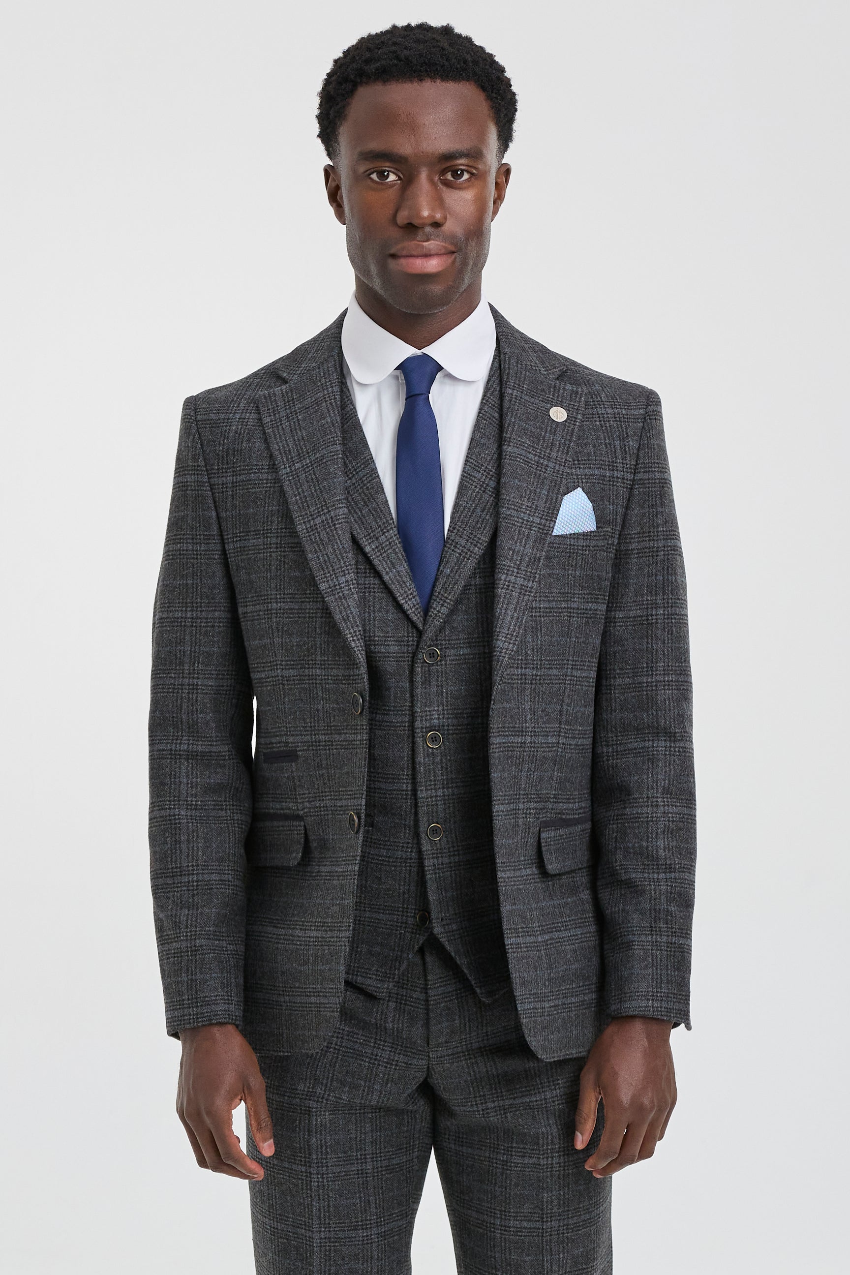 Hombre con traje gris a cuadros, corbata azul y pañuelo de bolsillo sobre fondo blanco.
