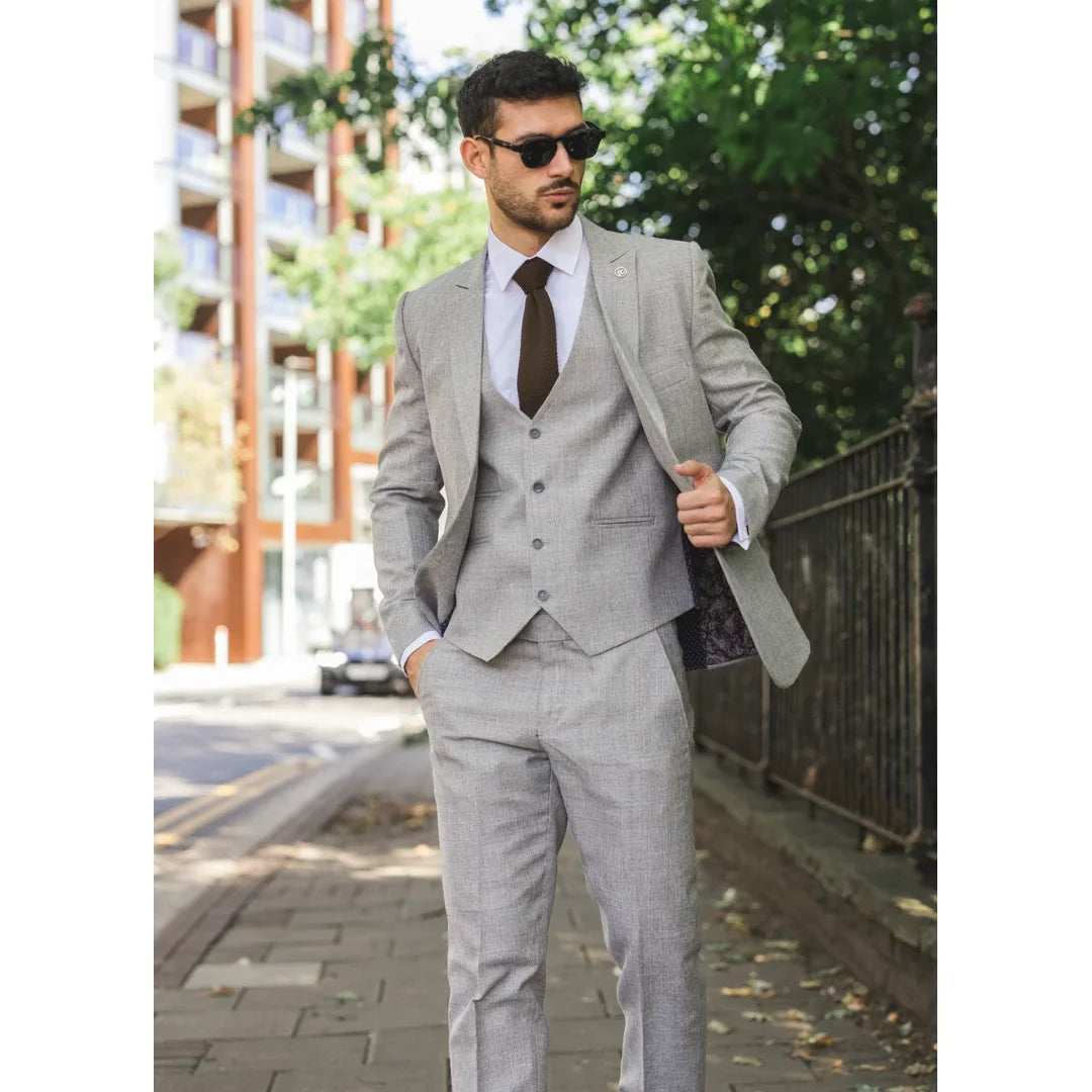 Hombre con traje gris caminando al aire libre con edificios y árboles al fondo.