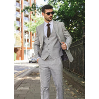 Hombre con traje gris caminando al aire libre con edificios y árboles al fondo.