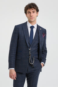 Hombre con traje a cuadros azul, camisa blanca y corbata azul sobre fondo gris claro.