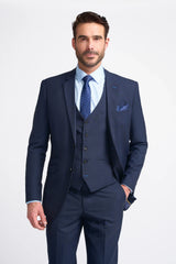 Hombre con traje azul marino, camisa blanca y corbata azul sobre fondo gris claro.