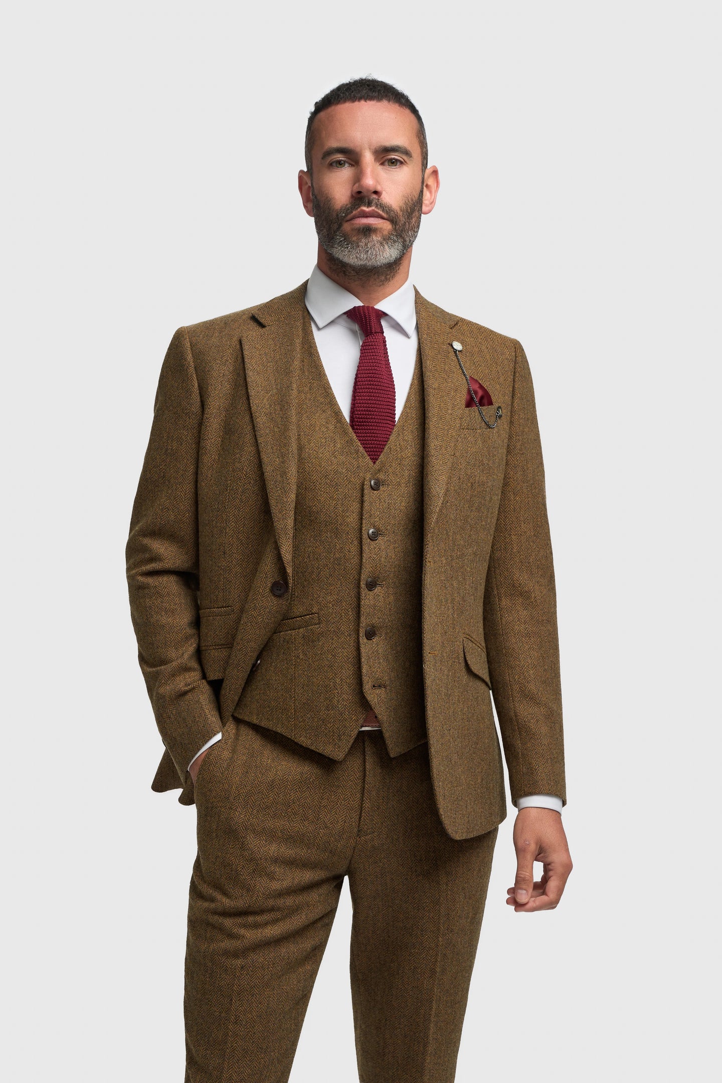 Hombre con traje marrón, corbata roja y pañuelo de bolsillo sobre fondo blanco.