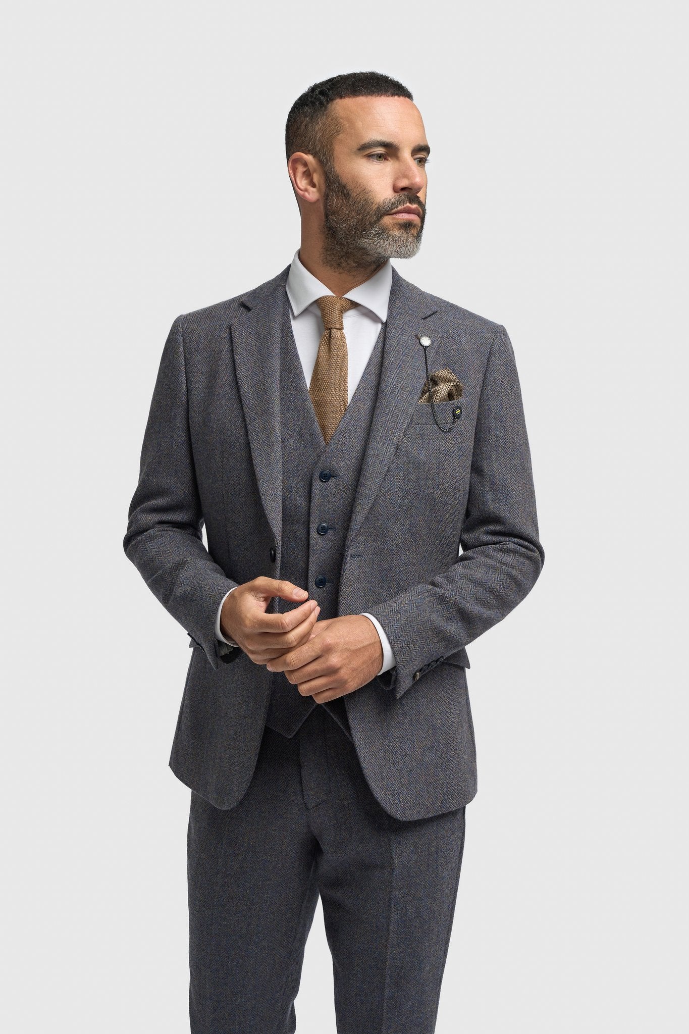 Hombre con traje gris, camisa blanca y corbata marrón sobre fondo gris claro.