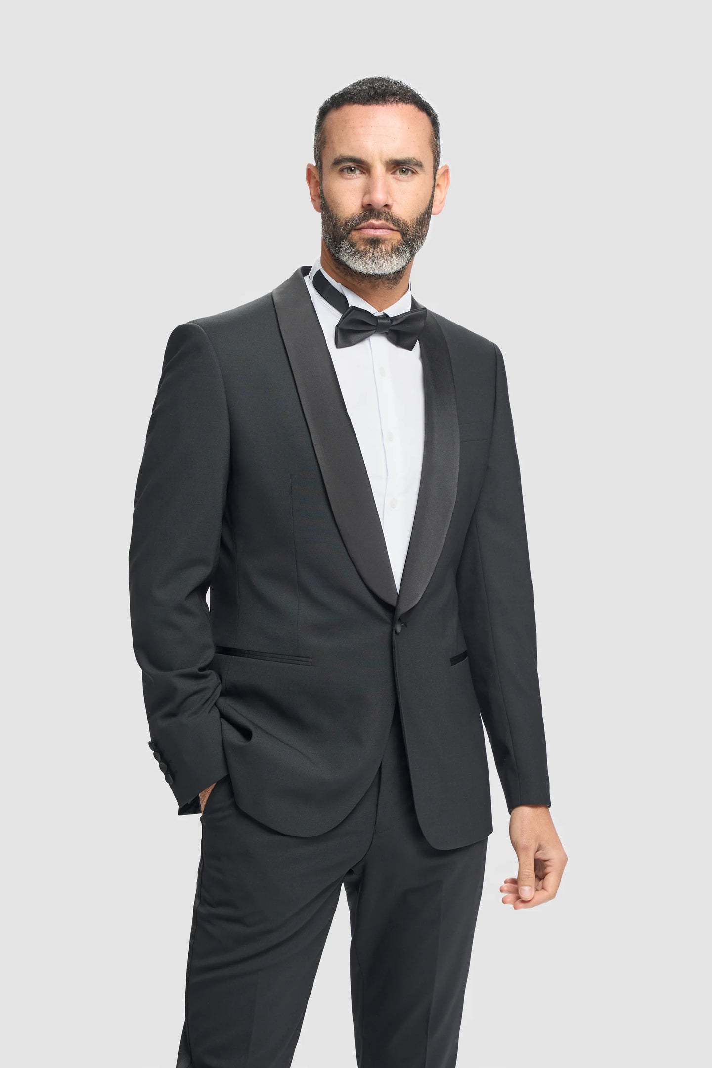 Hombre con esmoquin negro, camisa blanca y pajarita negra sobre un fondo gris claro.