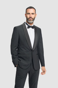 Hombre con esmoquin negro, camisa blanca y pajarita negra sobre un fondo gris claro.