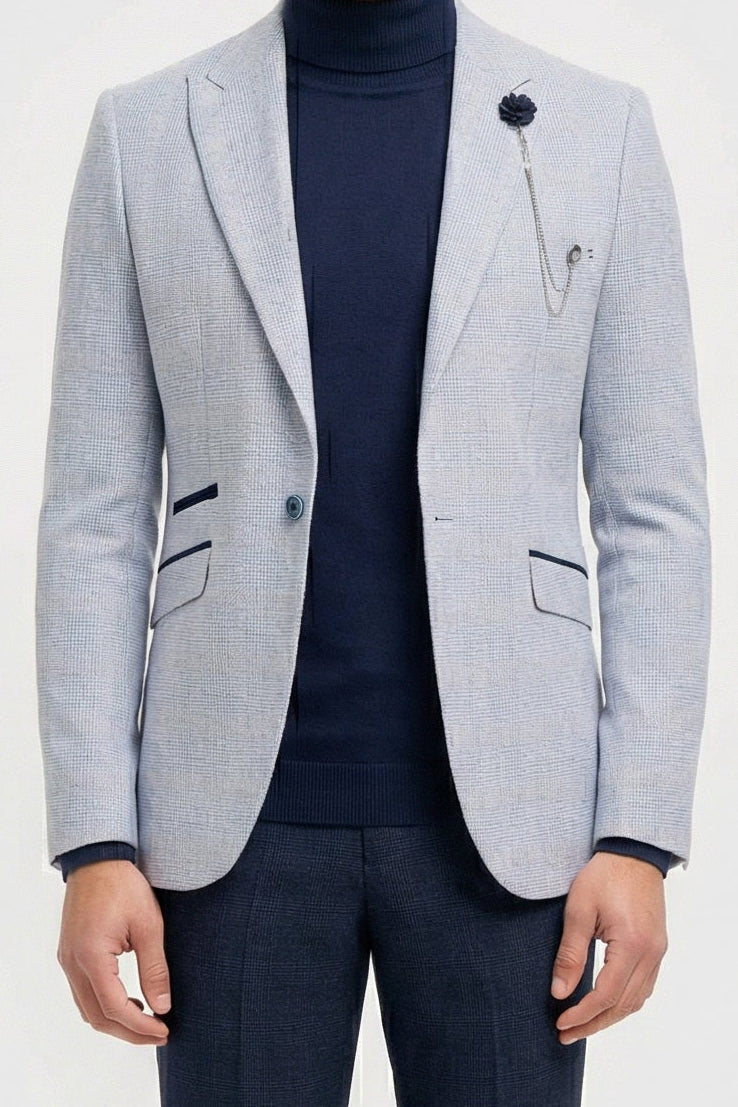 Hombre con chaqueta gris claro, jersey de cuello alto azul marino y pantalones oscuros sobre fondo blanco.