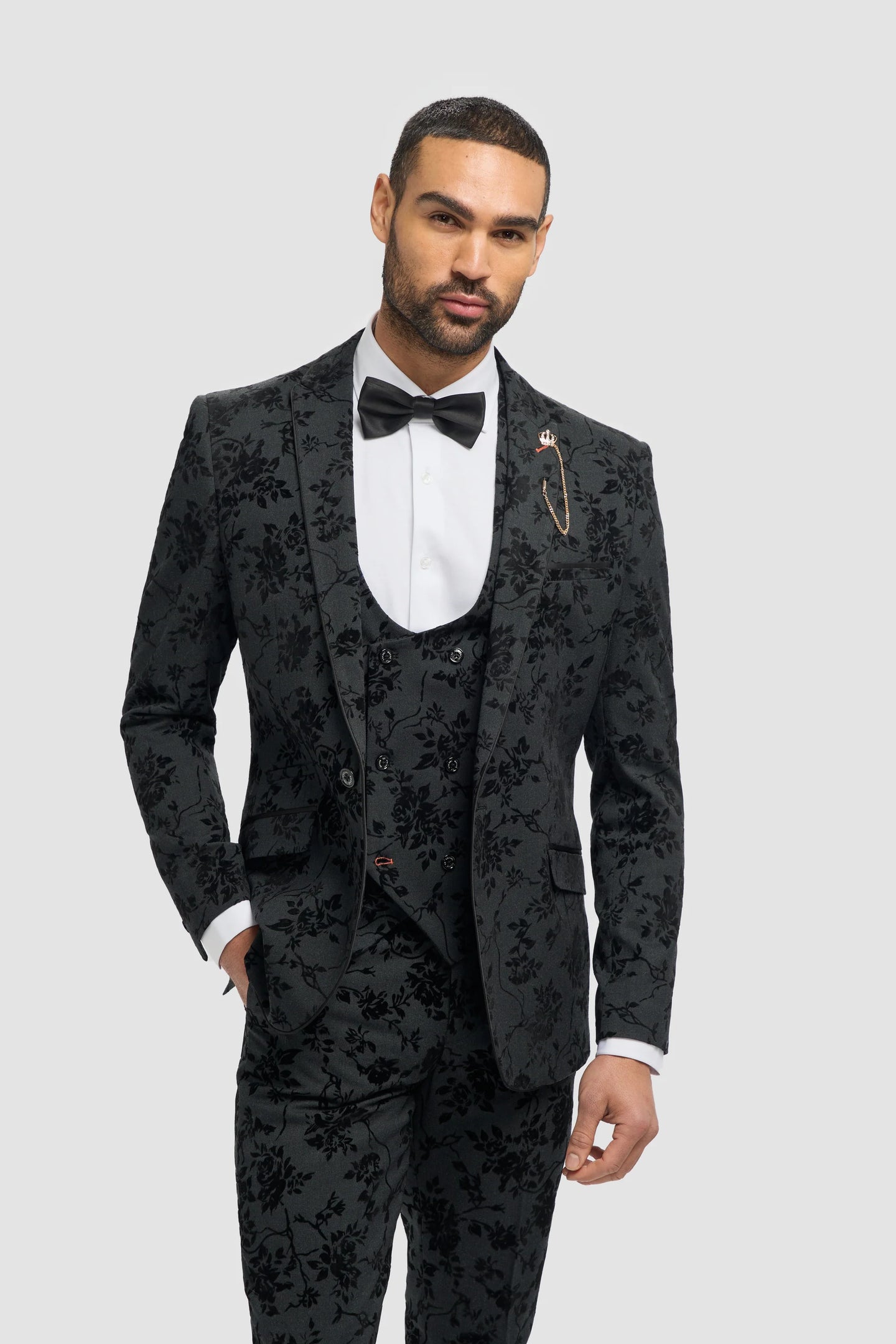 Hombre con traje negro estampado floral, camisa blanca y pajarita sobre fondo gris claro.